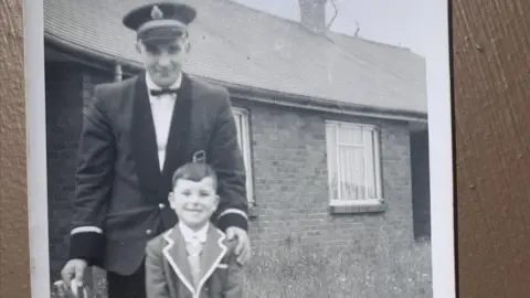 Steven Hickman A black and white image of a man standing with his arm around a young boy in front of a building. In his right hand he holds a brass instrument. Both are wearing smart clothes.