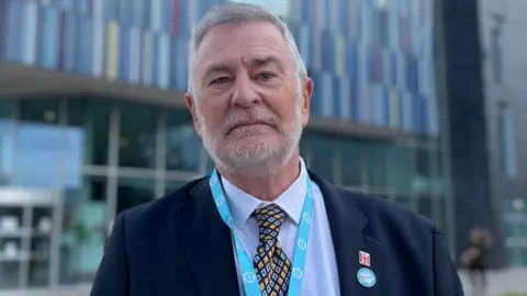 Guy Aston looks at the camera. He has short grey hair and a grey beard. He is wearing a dark suit, blue shirt and colourful tie. He has a Union Flag and Reform UK pin bad attached to his lapel.