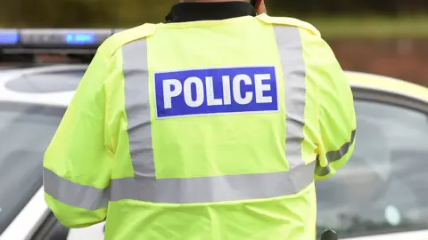A generic image of a police officer in a high-vis jacket with word 'Police' on the back and a police car in backgorund