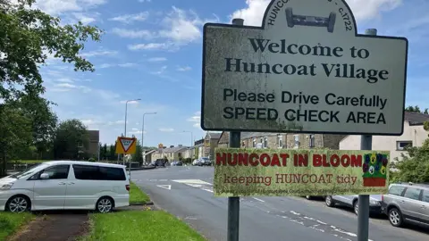 Dirty white sign saying 'Welcome to Huncoat Village' and a dirty yellow sign saying 'Huncoat in Bloom' on the road into the village on a sunny day.