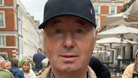 Ian Whitehead, a clean-shaven man with white hair, is pictured looking at the camera. He is wearing a black baseball cap. He appears to be sat in the outside area of a bar or restaurant and other groups of people can be seen sat around tables in the background. 