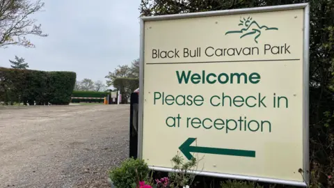 A large sign with green text which reads Black Bull Caravan Park Welcome Please check in at reception. Behind the sign is a gravel track and tall hedge.