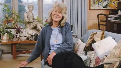 Getty Images Jilly Cooper sat on her couch smiling at the camera while wearing a dark blue sweater