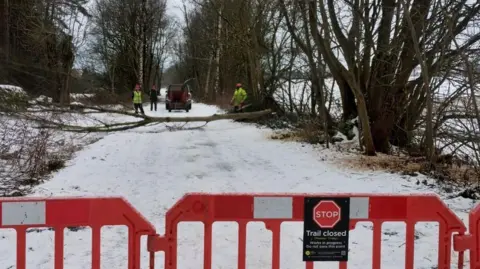 The Monsal Trail covered in snow with a red barrier and a "stop trail closed sign" at the front of the photo. Behind the signs, is a tree on the ground.