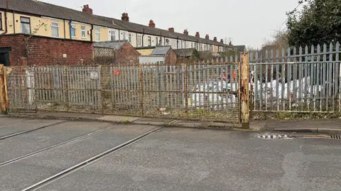 BBC/Paul O'Gorman A fenced-off area over the train tracks which cut across a suburban road. Bags of rubbish can be seen through the fence and to the left are the backs of a row of terraced houses.