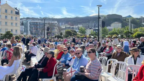 People at Liberation Square for Liberation Day Jersey from a previous year. Crowds have filled the square. They are sat on white plastic chairs. It's a sunny day with blue skies. Buildings can be seen in the background. People are wearing hats and sunglasses and some are holding the Jersey flag.