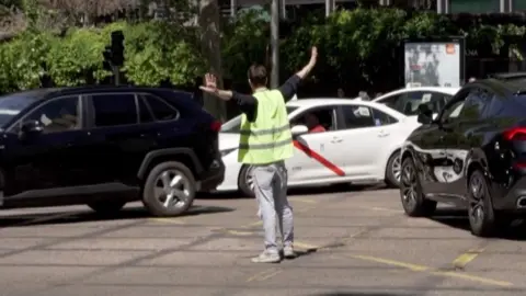 A man in a high-visibility jacket is directing traffic in the middle of the road, cars are passing him