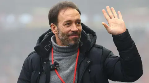 Rex Features Iñigo Calderón in a black coat and grey turtleneck jumper, waving.