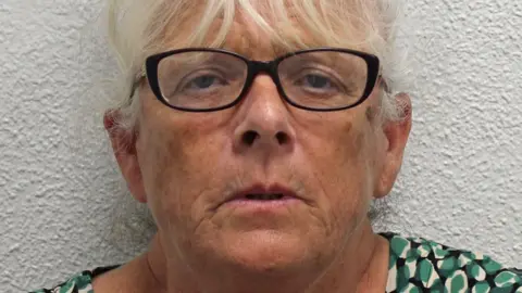 Deborah Mason, a woman with white hair and wearing dark rimmed glasses. She is standing against a white background and wearing a green, white and black patterned top