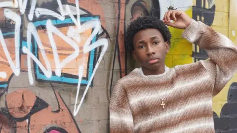A boy with black curly hair poses against a graffitied wall