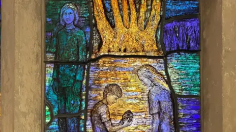 Image of a stained glass window depicting a boy who is kneeling down looking at a shell. A young girl is standing over him while a second girl looks on. 