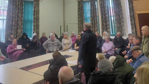 David Torrance A room with a high ceiling and long curtains. A man is standing in the centre on a microphone surrounded by lots of residents.