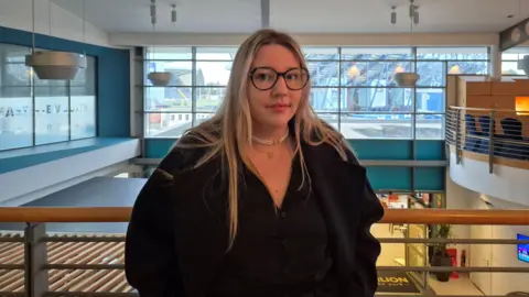 Lisa Beacham standing against a railing on the upper floor of a building. She has blonde hair past her shoulders, glasses and is wearing a black coat and two necklaces.