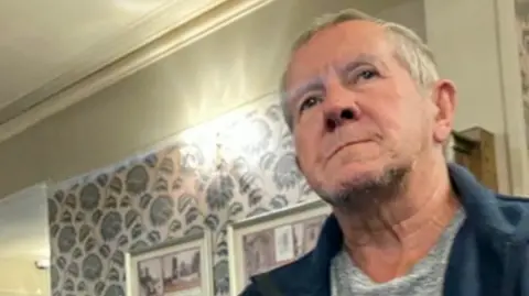 Contributed An older man with short grey hair wearing a light grey top with a darker blue top over it. He is in a room with patterned wallpaper and bright lights