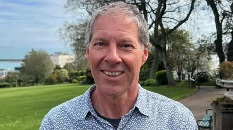 A balding grey haired man smiles at the camera, wearing a patterned blue, gold and yellow shirt. Behind him lays greenery and trees in a park setting.