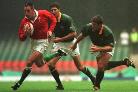 Getty Images Mark Taylor charges towards the tryline, ball-in-hand, with a determined look on his face. Chasing him are two South African players, dressed in white shorts and green shirt with gold collar 