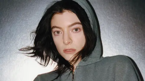 Lorde. She is pictured against a silver background wearing a grey hoodie with the hood up. She has long dark hair which is peeking out from underneath the hood.