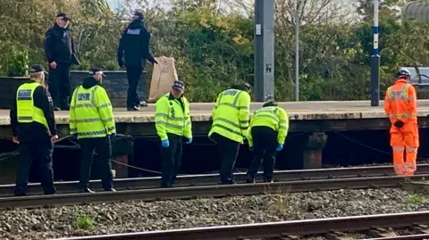 Shaun Whitmore/BBC A group of transport police officers walk along the track, looking closely for clues. They wear high-vis jackets over black hoodies and black trousers., One railway worker dressed in orange walks ahead of them.