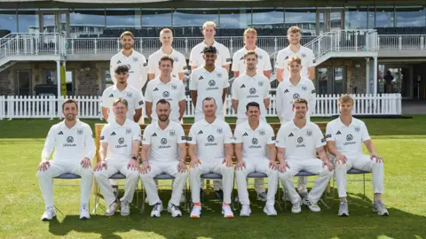 Getty Images A group shot of the Gloucestershire cricketers. They are posed in three rows, wearing their right uniforms. The front row are sat down with their hands on their knees, and the two rows in the back are standing with their hands clasped behind their backs. It is a sunny day and the club building is visible in the background, with lots of glass windows and white picket fences.