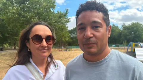 A couple stand in front of a parched park with the paddling pool behind. The woman is wearing a white shirt with large brown sunglasses. The man is wearing a grey t-shirt.