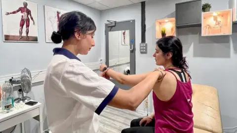 Sonia Kumar MP A woman with tied-back black hair in a white medical top holding the arm of another woman with dark hair and a pink vest top on a couch in a white-walled room