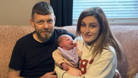 Family handout Photo of a man and woman sitting on a sofa together. The woman is holding a young baby in her arms, the man has one of his arms around the woman.