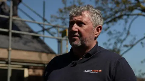 A man with grey hair and a black fleece stands in front of a house with solar panels.
