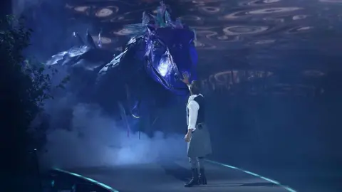 Getty Images The actor SamHeughan stands onstage, wearing a kilt, and with his back to the camera. He is looking at a giant Loch Ness monster prop that is emerging on stage. It is scaled and coloured purple.