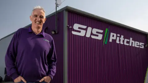 SIS Pitches George Mullen standing outside SIS Pitches in Maryport. He has short white hair and is wearing a purple jumper with the company's logo. He is standing in front of the factory, which has purple cladding and the company name on the side.
