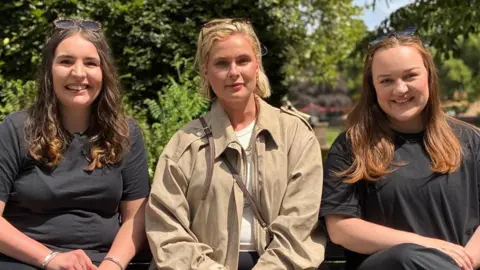 All three women in their late twenties and early thirties are sat on a bench in a park. Cari is on the left wearing a black outfit. Mererid is in the middle wearing a light beige coat and Nia is on the right in a black outfit. 