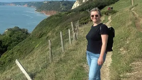 Lizzy Willmington Lizzy Willmington smiling at the camera while walking on a coastal path overlooking the sea. She is wearing a black t-shirt, blue jeans and sunglasses with her hair tied back and is carrying a backpack. 