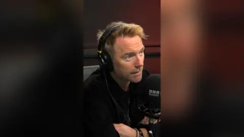 Ronan Keating sat in a radio studio with black headphones on and a black mic in front of him.