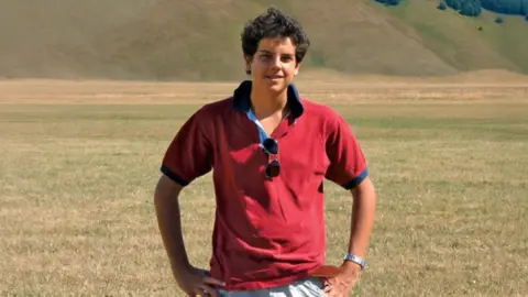 BBC A boy with dark curly hair in a red polo shit stands, smiling at the camera, with his hands on his hips, in front of a field and hills