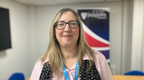 A woman with dark blonde hair and black glasses is wearing a pink shirt, blue lanyard and a black scarf with white spots on it. She is smiling at the camera.