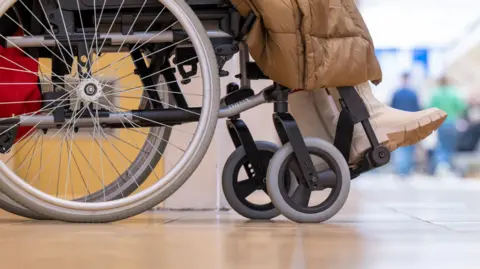 Stock image of wheelchair and photo of someone's feet with white shoes and brown jacket overhanging. 