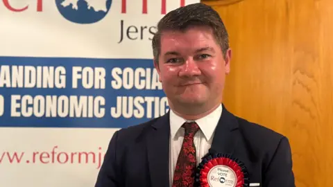 Sam Mezec wearing a navy blue blazer, white top and red tie. He is wearing a Reform badge. He is smiling at the camera. 