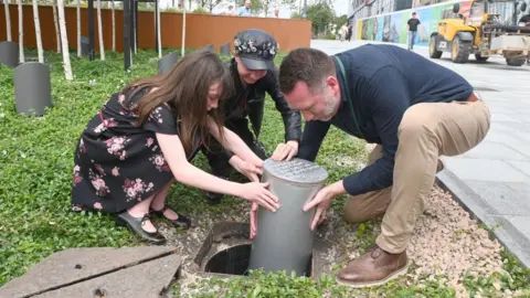 Three people on Armada Way in Plymouth buried the time capsule which will not be opened until 2047. They are dropping the metal cylinder into place. 