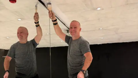 Josh Baines A man stood in front of a mirror holding a paint brush up to the ceiling 