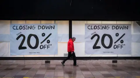 Getty Images A man in a red coat walks past a shopfront with windows displaying large posters, the text on the posters reads: "CLOSING DOWN, EXTRA 20% OFF"