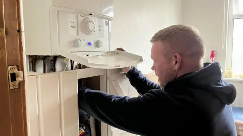 James Hunt, in blue hoody with short sandy hair, working on a domestic heating boiler in a bathroom