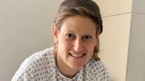 A woman looks at the camera. She has dark blonde hair, pulled behind her head and wears a hospital gown of white with a dark diamond pattern on it. 