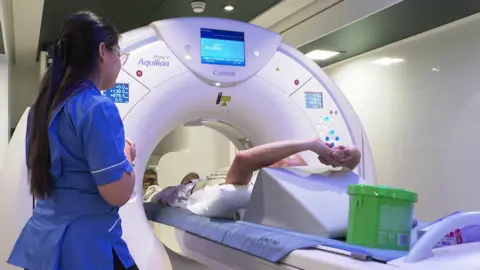 A man is lying on a bed facing away from the camera with his arms raised above his head. His body is lying under a large hoop-shape machine that has a series of buttons on it. A nurse is standing next to him. 