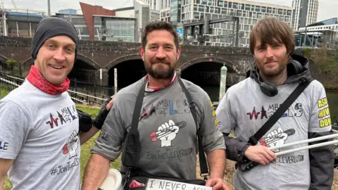 Three men wearing grey tshirts with Wambles of Wales on the fronts of them. The one to the left is wearing a hat and red neckerchief. The man in the middle has dark hair and a beard. The one to the right have dark hair, a beard and he's holding a litter picker. They're standing alongside the Taff.
