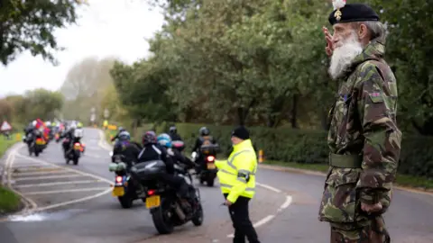 PA Media A large group of motorbikes on a road. A steward in a green high visibility jacket is on the road. At the front of the image a man wearing a uniform with a camouflage pattern who is saluting the motorcyclists. He is wearing a beret and has a grey beard and dark grey hair. 