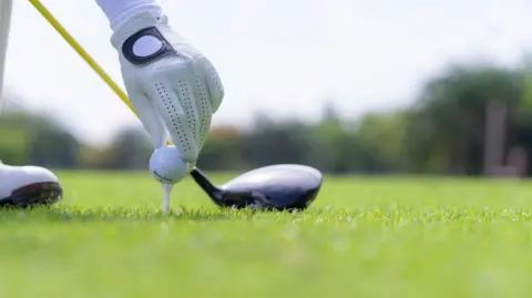 A gloved hand can be seen placing a white golf ball on a golf tee in the grass; a golf club is visible behind this and the corner of a white shoe can also be seen in the frame. In the background are blurred trees.