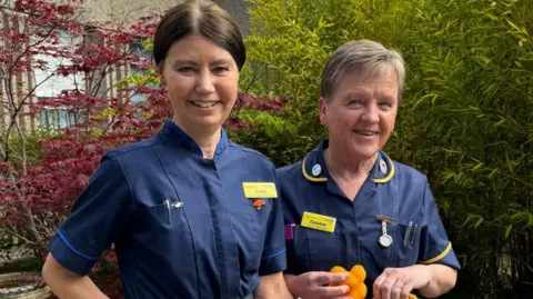 SATH Two women in dark blue hospital uniforms, one with dark hair and the other with blonde hair. One of them is holding a small orange knitted object