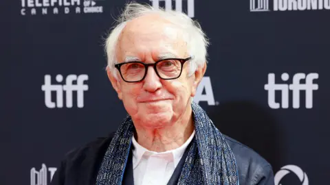 Getty Images Headshot of Pryce against a photo call background. He wears dark framed glasses. He wears a dark blue shirt jacket over a white shirt. He wears a blue patterned scarf over his shoulders