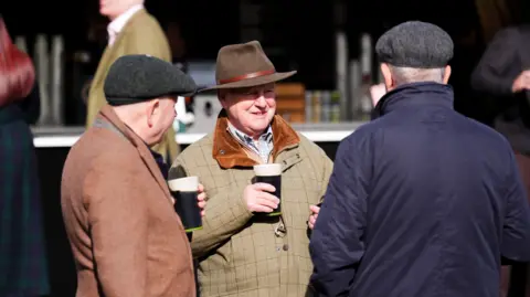 PA Three men wearing jackets and hats gather near an outdoor bar, holding pints of Guinness, smiling and chatting with each other. It is a bright sunny day but still looks cold.