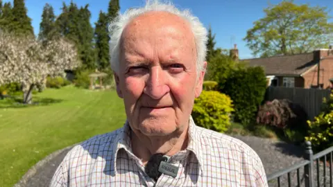 A man is standing in a garden on a sunny day. There is a lawn behind him as well as trees. He has grey hair and is wearing a checked shirt. There is a bungalow to the right of the image.