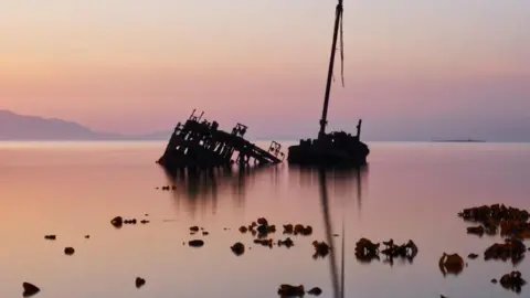 Sandy McGrattan The rusted remains of a shipwreck lean in calm, glassy water at sunset, with soft pink and orange light reflecting across the scene.